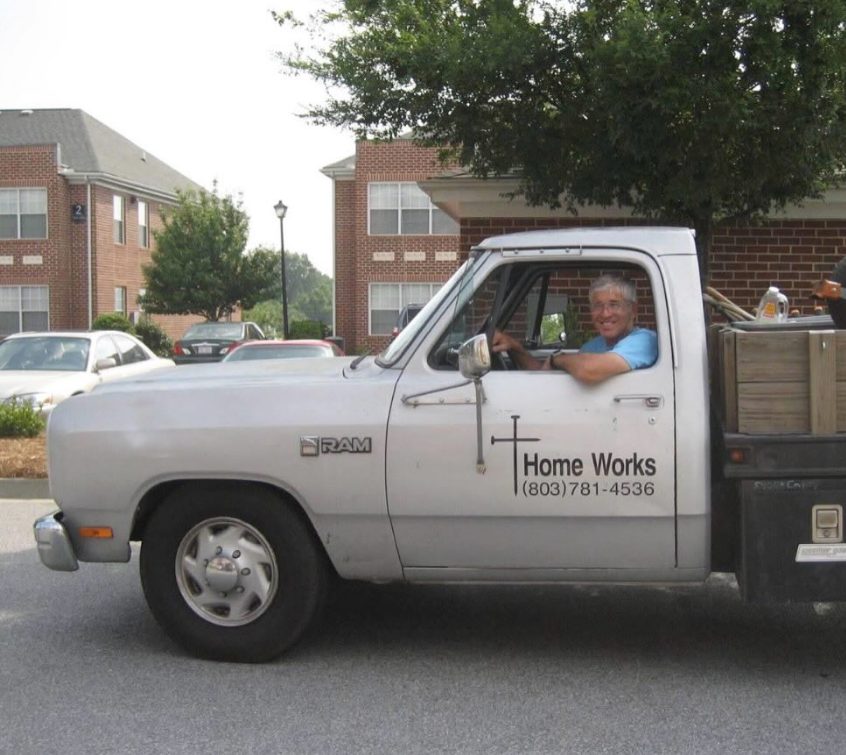 Darryl K. Williams smiling from the driver's seat of a white Home Works of America pickup truck, with a cross and the Home Works name on the door